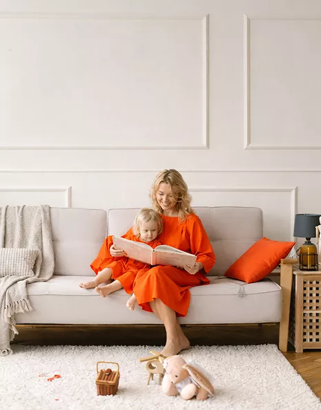 A mother in an orange dress sitting on a beige sofa reading a book with her toddler in a bright, elegantly styled living room.