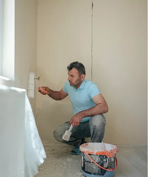 Painter crouching to apply filler near a skirting board with a small roller and a bucket of compound beside him.