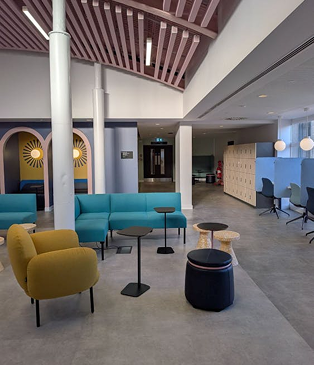 Modern office lounge with teal and mustard modular sofas, slatted ceiling detail, white columns, and colourful accent walls.