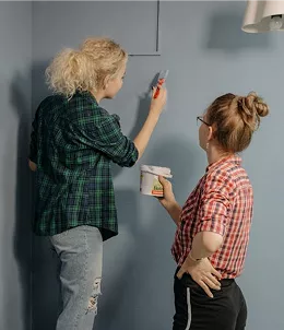 Two women painting a grey wall together, one on a step holding a brush and the other holding an open paint tin