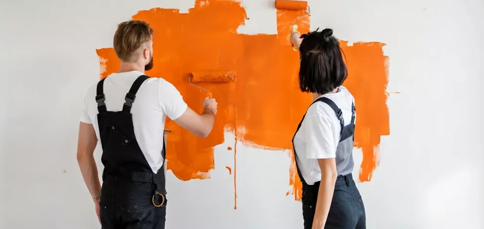 Two painters in black overalls rolling bright orange paint onto a white wall, viewed from behind.