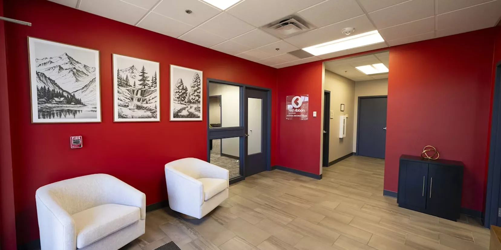Office reception area with bold red walls, two white armchairs, nature-themed framed art, and a hallway leading to private rooms.