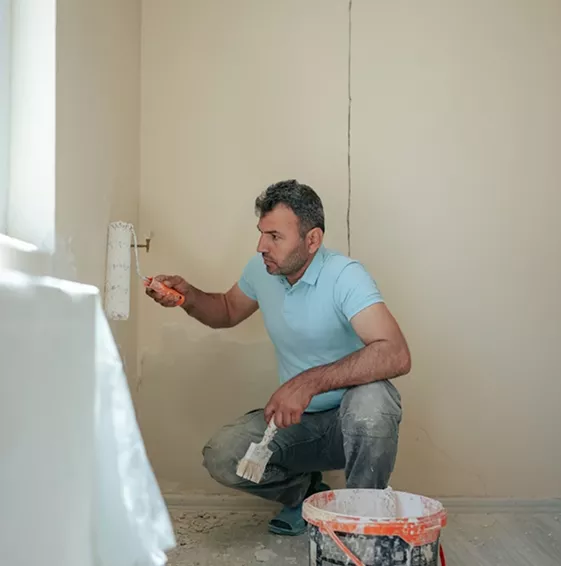Painter crouching to apply filler near a skirting board with a small roller and a bucket of compound beside him.