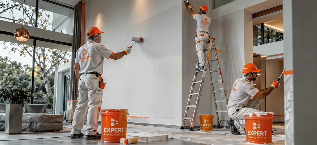 Three uniformed painters working on a white interior wall — one rolling, one on a ladder, one cutting in at the base.