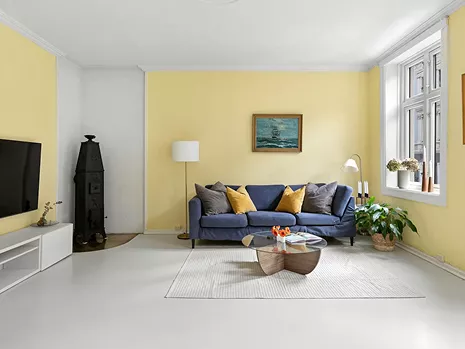 Bright living room with soft yellow walls, a navy blue sofa with mustard cushions, and a large window letting in natural light.