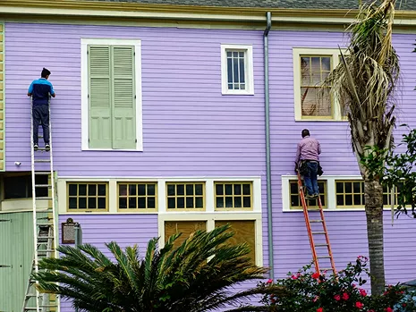 Two painters on ladders painting the upper storey of a purple wooden house exterior with white trim and shutters.
