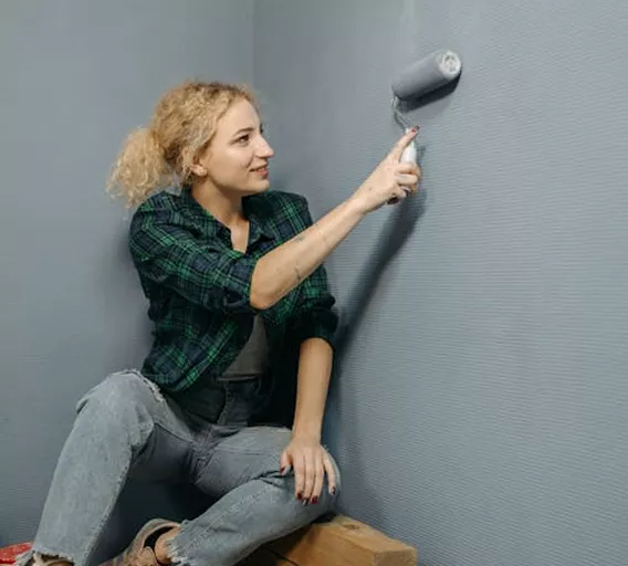 Young woman in a plaid shirt smiling while rolling grey paint onto a corner wall in a room under renovation.