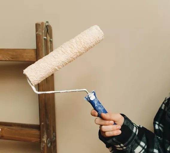 Close-up of a hand holding a paint roller beside a wooden ladder against a beige wall.
