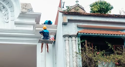 Two workers on a suspended platform painting the white ornate exterior walls and cornices of a heritage-style building