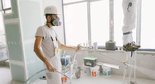 Painter in a respirator mask and hard hat using a spray gun to apply paint inside a building under construction.