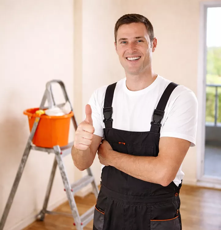 Smiling male painter in dark overalls giving a thumbs up inside a room with a ladder and paint bucket behind him.
