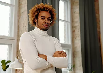 Young man with curly hair and glasses in a white turtleneck, arms crossed, standing in a bright loft-style room.