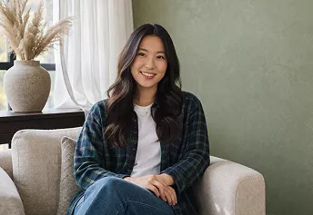 Young woman in a plaid shirt sitting on a sofa and smiling, with a sage green wall and dried flowers in the background.