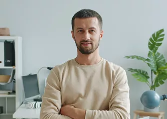 Man in a beige sweatshirt with arms crossed, smiling in a casual office with a plant and desk behind him.
