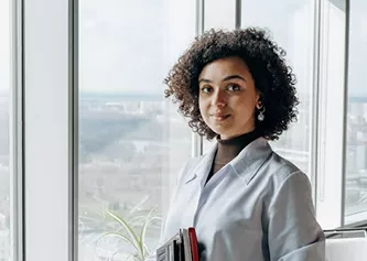 Professional woman with curly hair holding notebooks, standing by large windows with a city view behind her