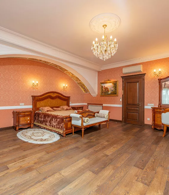 Grand classic bedroom with pink damask wallpaper, an ornate wooden bed, crystal chandelier, and arched alcove detail.