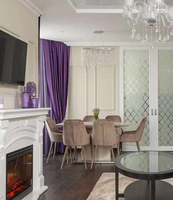 Elegant dining room with cream walls, purple drapes, a crystal chandelier, velvet chairs, and a white decorative fireplace.