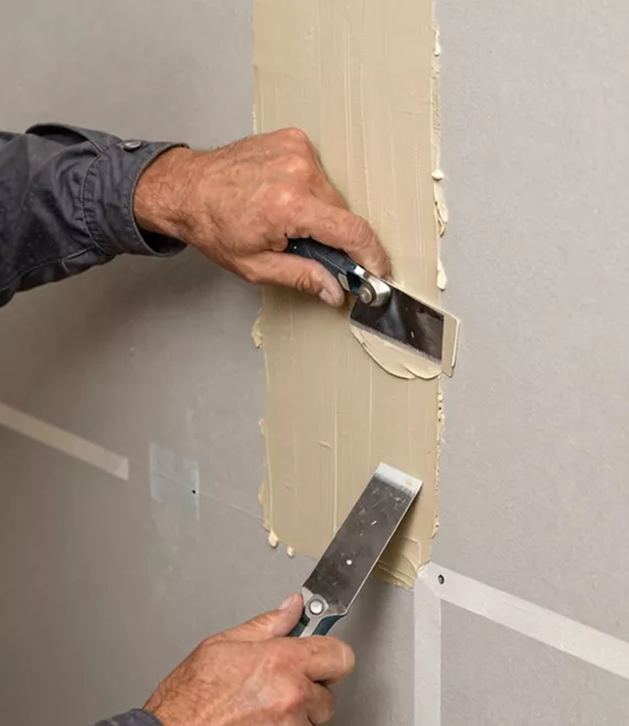 Close-up of two hands using putty knives to apply and smooth filler compound onto a drywall seam.
