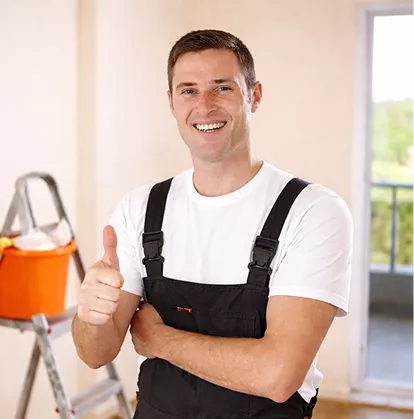 Smiling male painter in dark overalls giving a thumbs up inside a room with a ladder and paint bucket behind him.
