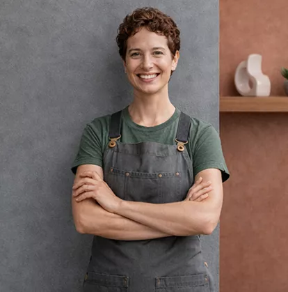 Woman in a grey apron and green t-shirt smiling with arms crossed against a textured grey wall.