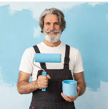 Senior male painter with grey beard holding a blue paint roller and tin, standing in front of a freshly painted blue wall.