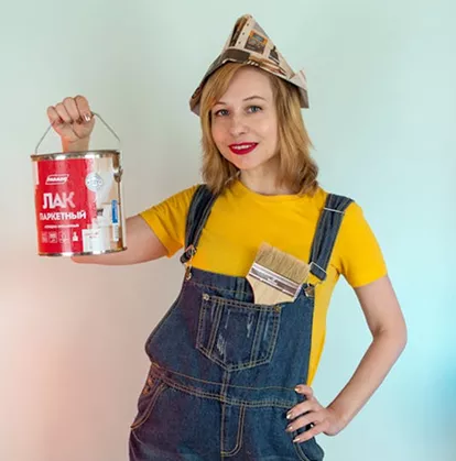 Smiling woman in denim overalls and a yellow top holding a paint can and brush, wearing a paper hat.