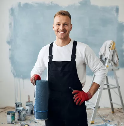 Male painter in black overalls and red gloves holding a blue paint roller, standing in front of a freshly painted wall.