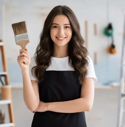 Young woman in a black apron smiling and holding a large paintbrush in a bright workshop setting.