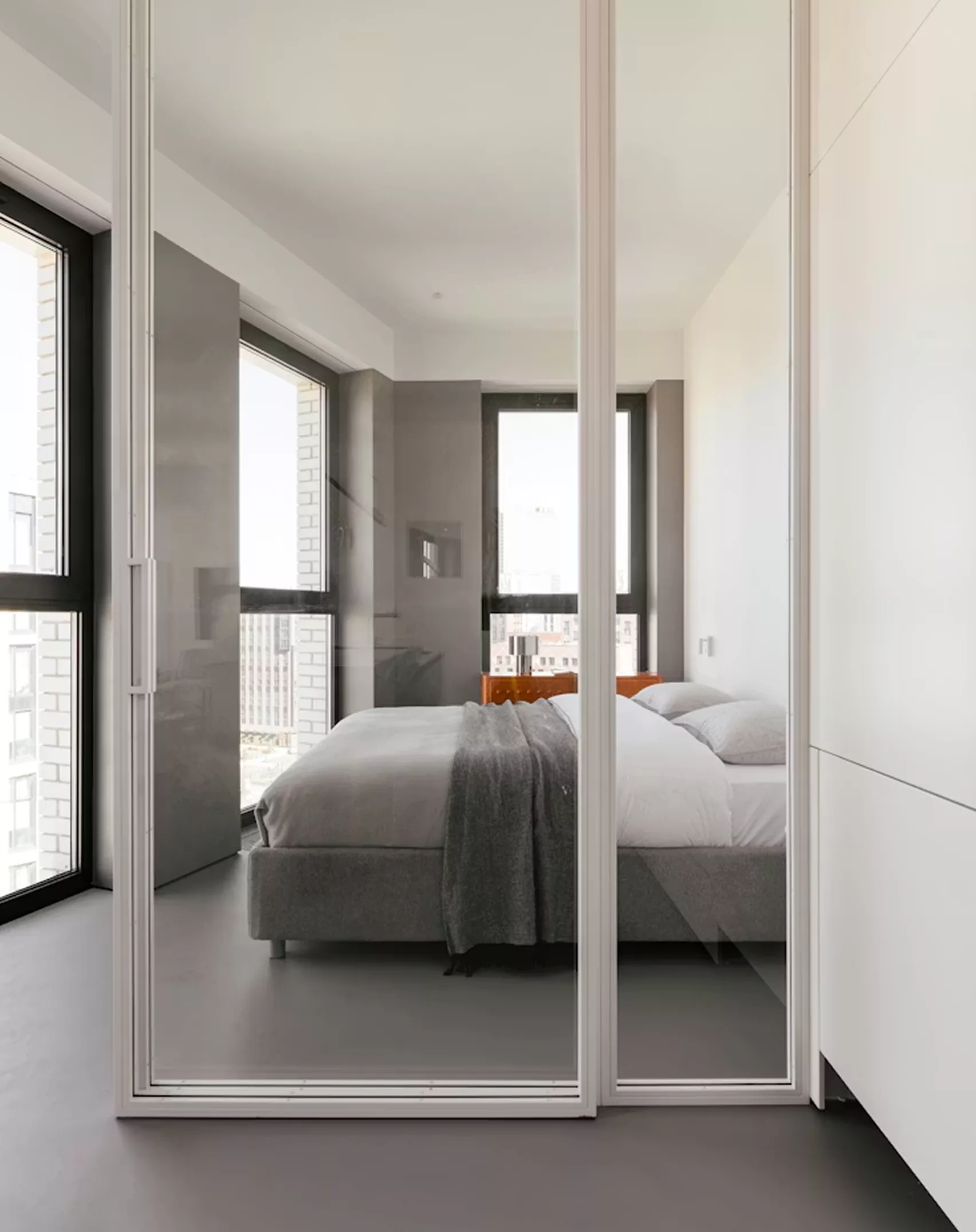Minimalist bedroom reflected in tall mirrored wardrobe doors, with a grey upholstered bed and city views through black-framed windows.