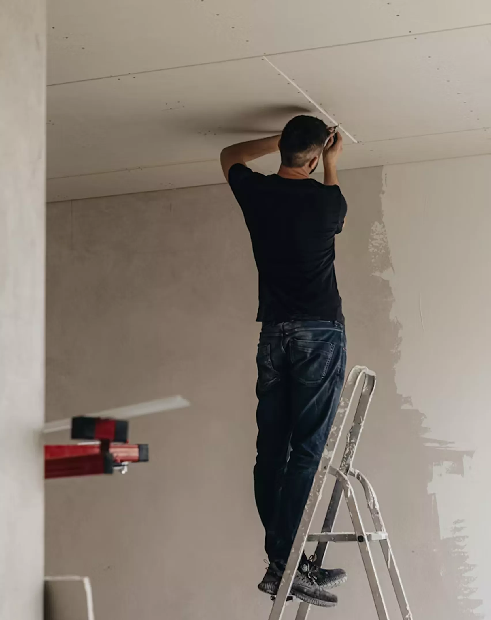 Worker standing on a ladder applying plaster to the ceiling junction of a room under renovation.