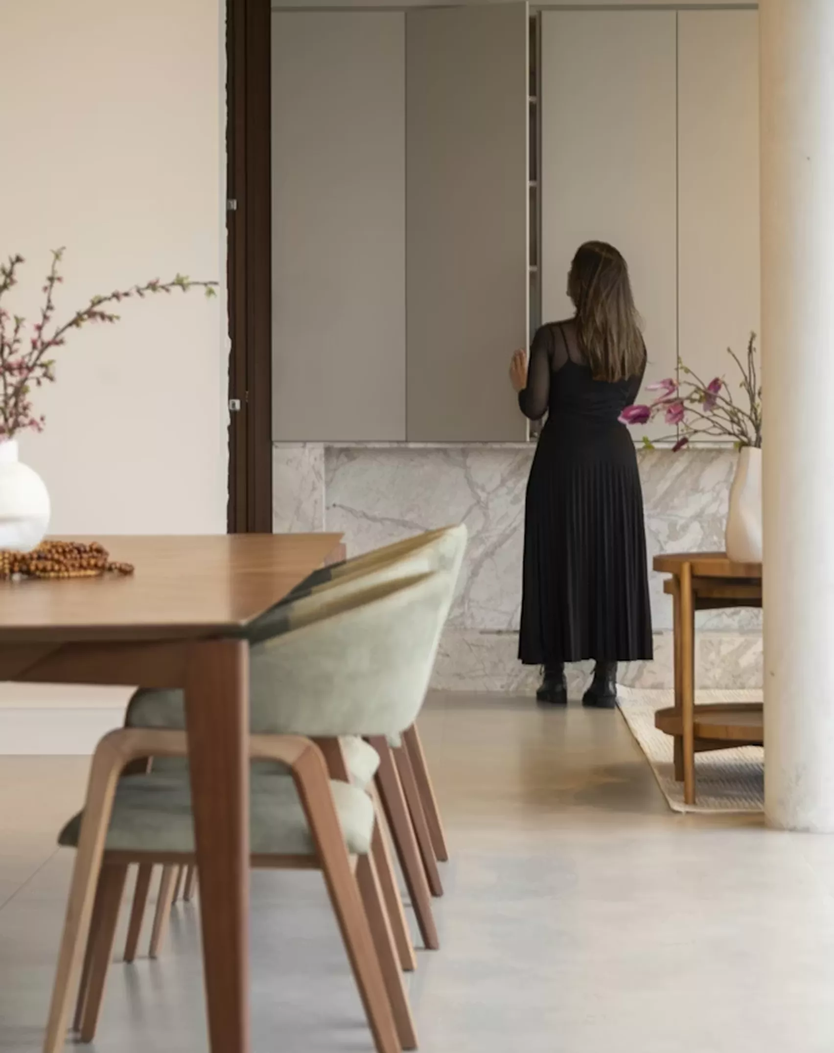 Woman in black standing at a marble console in a serene open-plan interior with a walnut dining table and floral stems in the foreground.