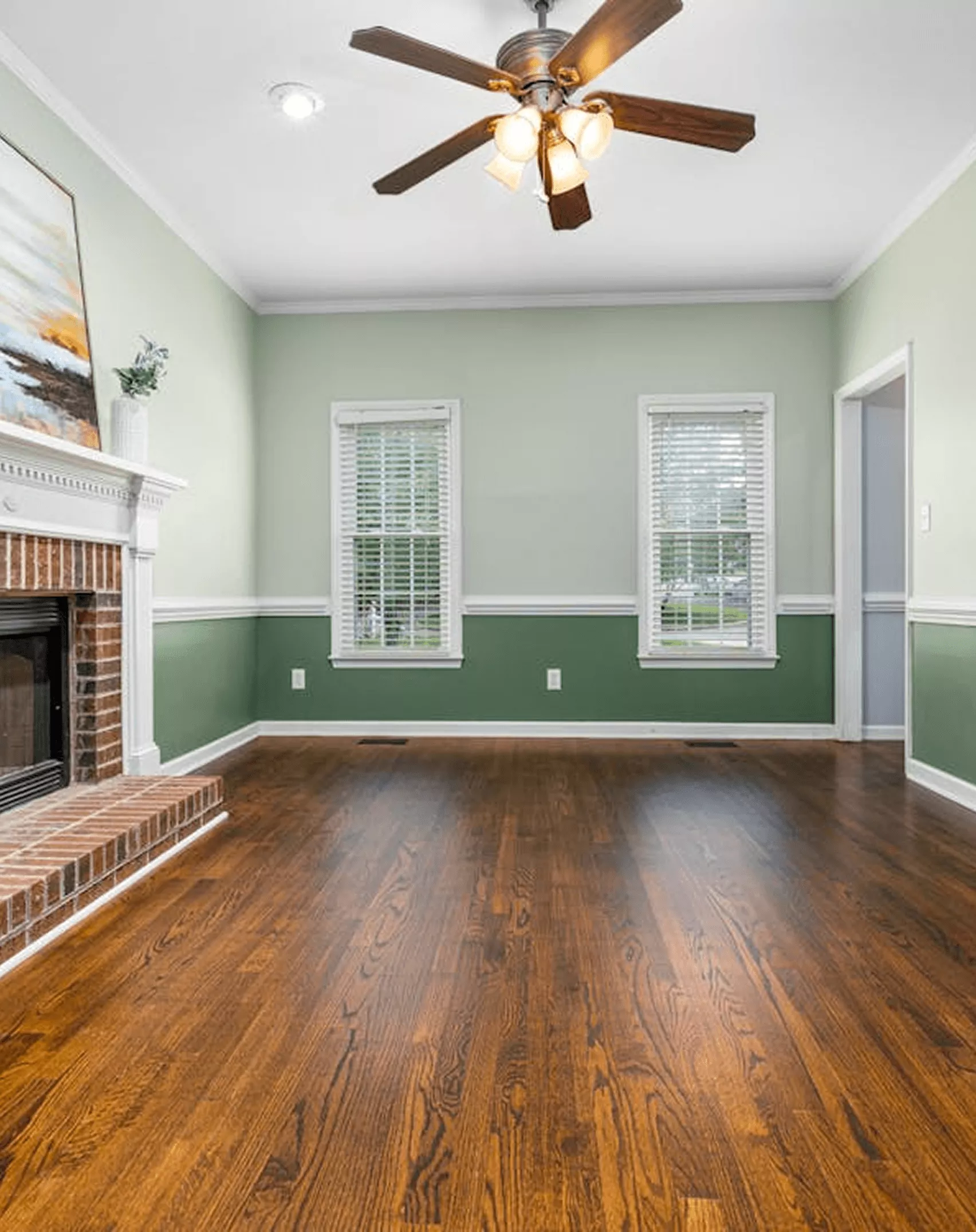 Empty living room with two-tone green painted walls, white trim, a brick fireplace, and polished hardwood floors.