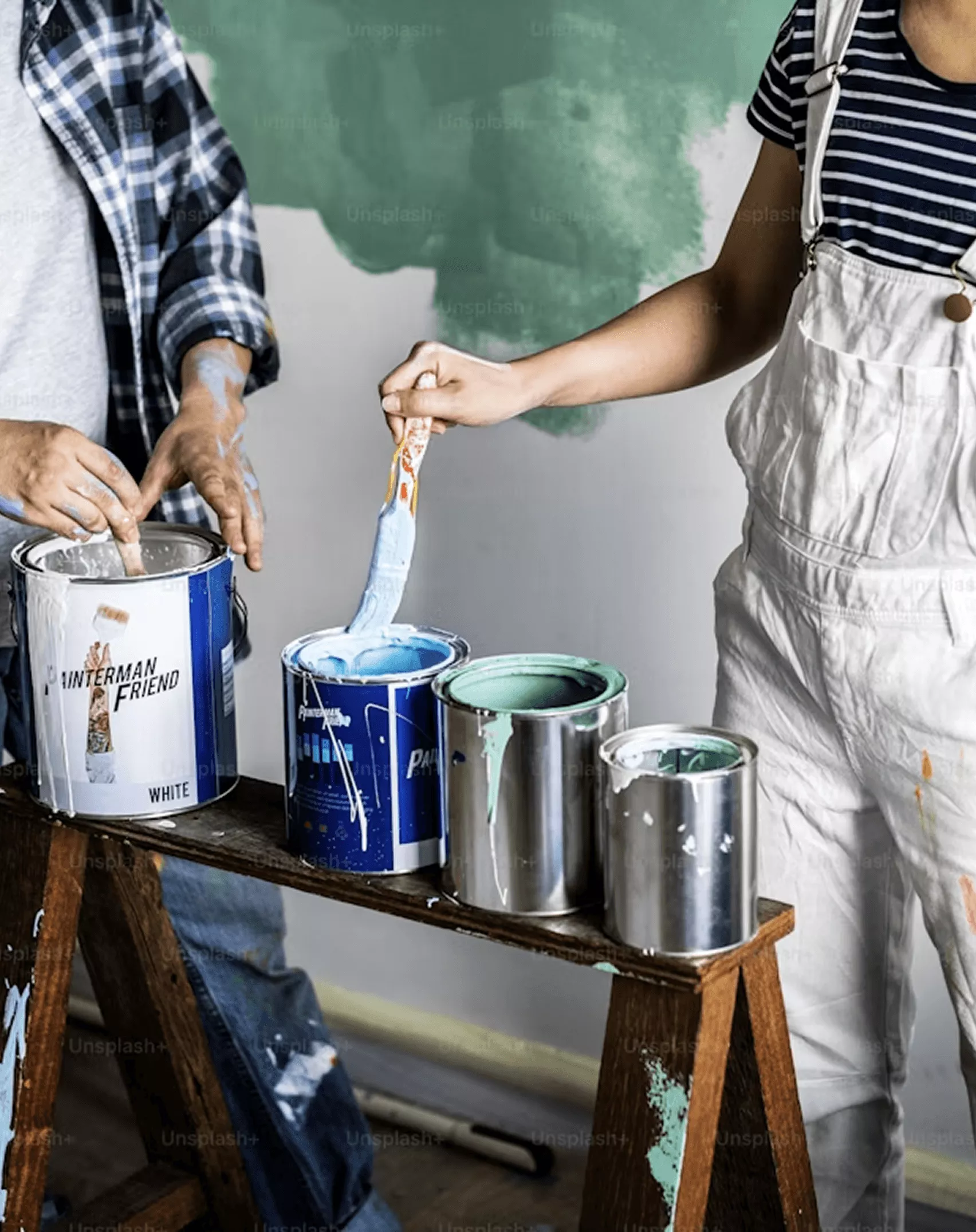Two painters mixing blue and white paint from open tins on a wooden sawhorse bench in front of a partially painted wall.